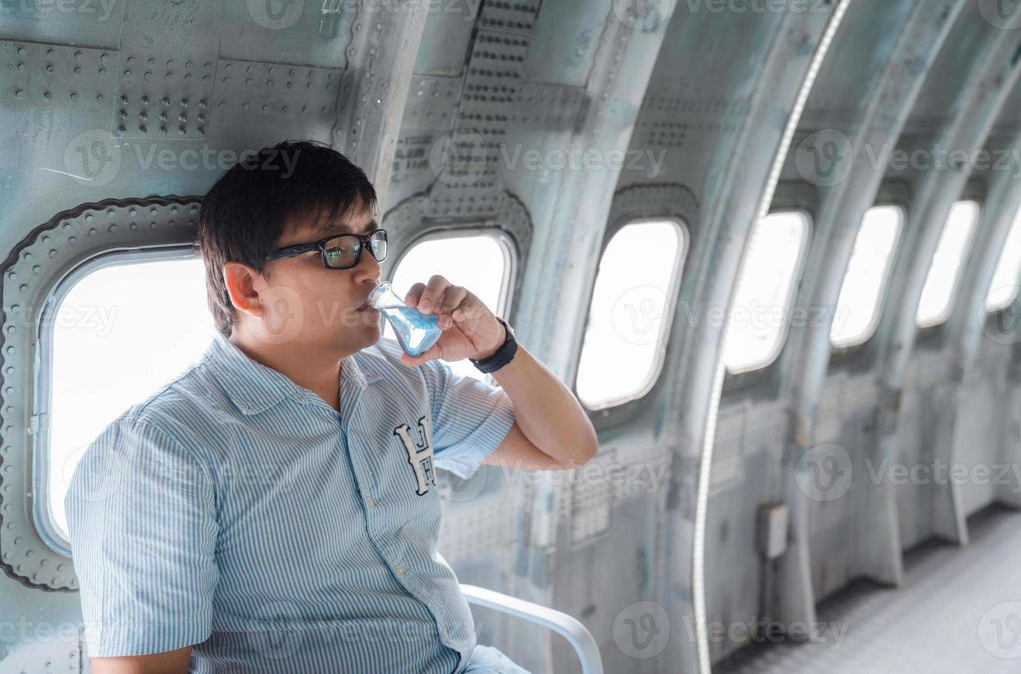 paunchy obese Asian man drink blue liquid from Erlenmeyer flask bottle on steel airplane near window photo
