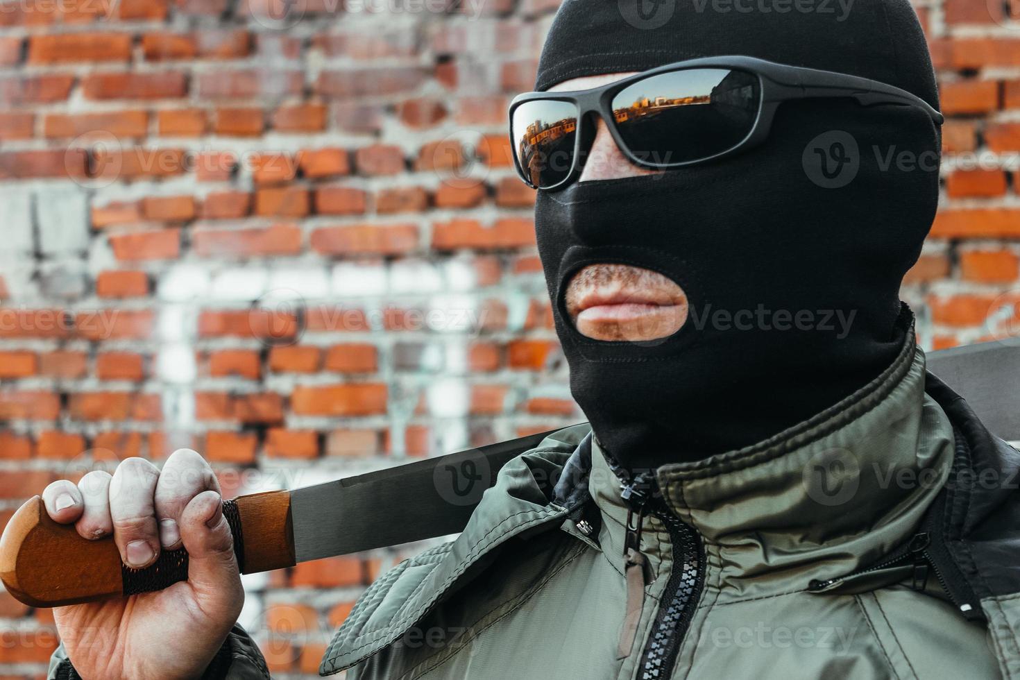 Commando in a mask and glasses with a machete in his hand against a brick wall photo