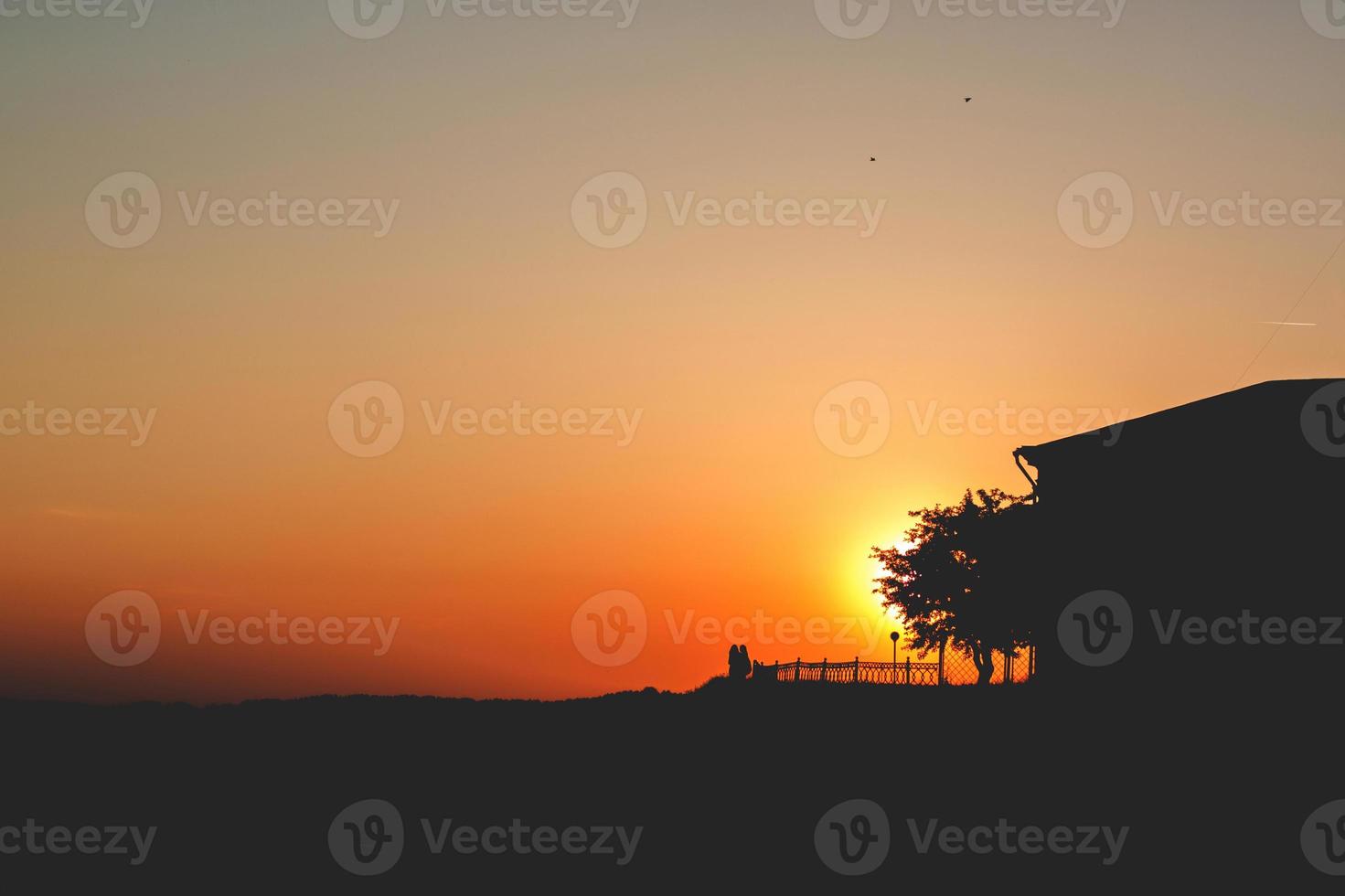 puesta de sol. dos personas se paran en una colina con un árbol y una casa puesta de sol foto