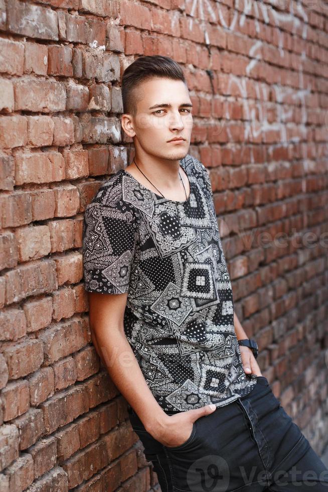 un joven hipster con una camisa negra parado cerca de una pared de ladrillo rojo con graffiti. foto