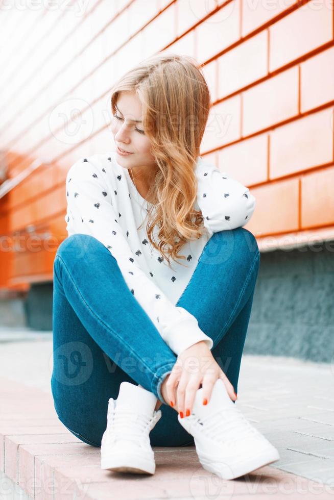 Beautiful woman in a white sweater, jeans and white sneakers sitting on ...