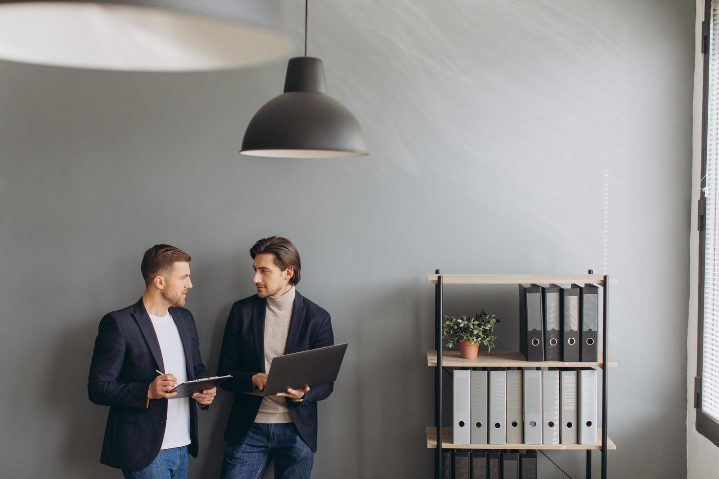 Young business men standing together holding a laptop in the modern office, discussing photo