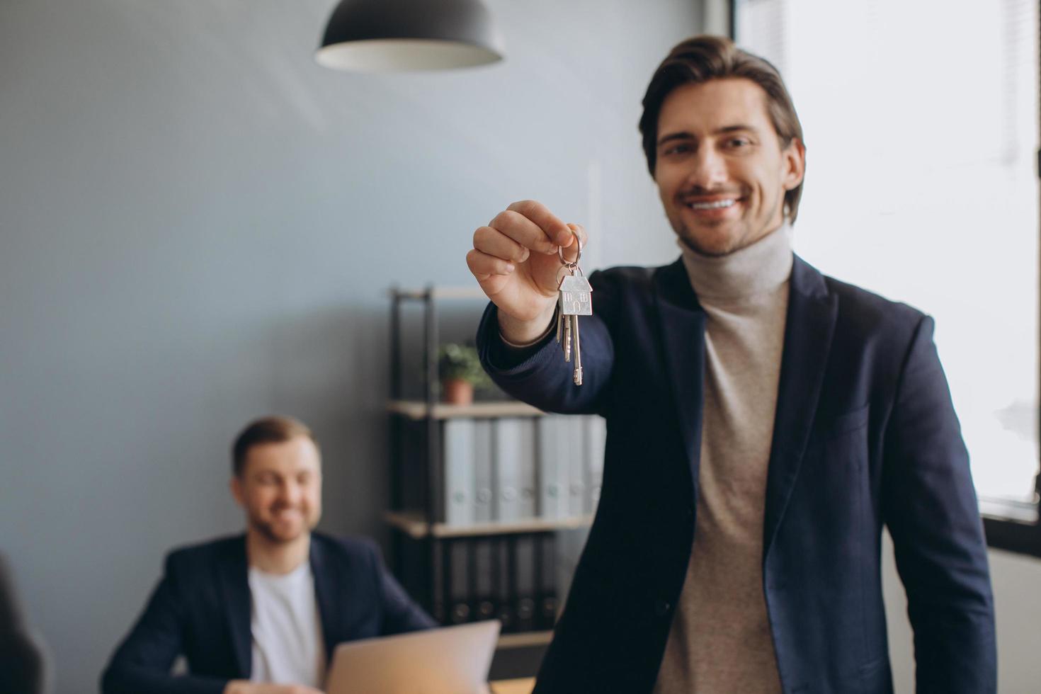 Smiling young modern man in a suit holds the keys to the bought apartment on the background of the realtor and office photo