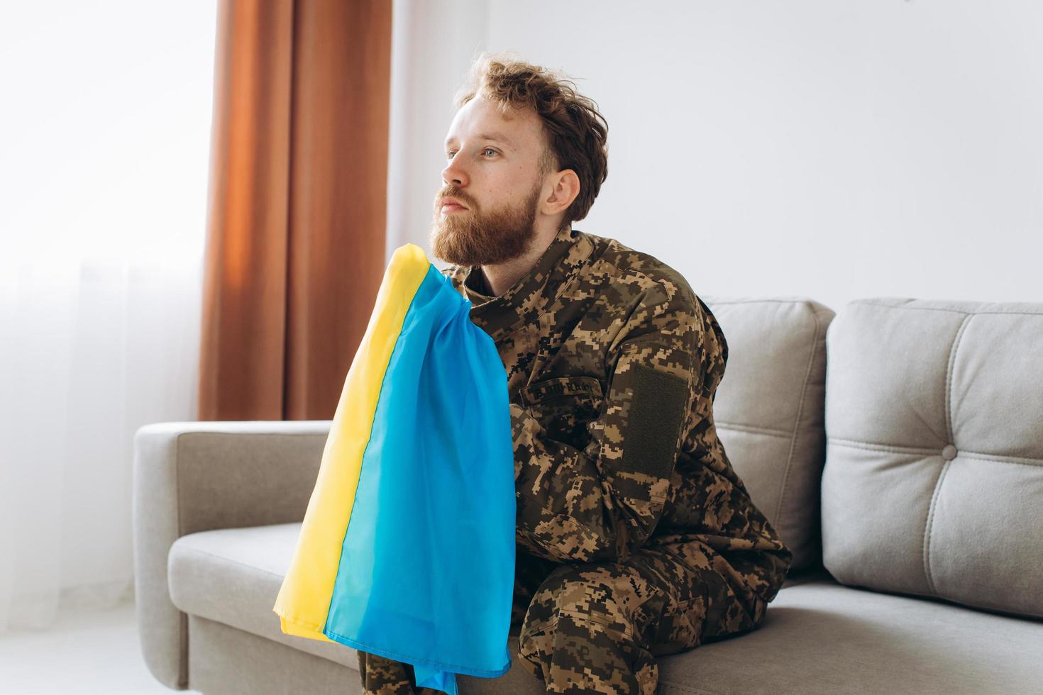 Portrait of an emotional young Ukrainian patriot soldier in military uniform sitting on the office on the couch holding a yellow and blue flag. photo