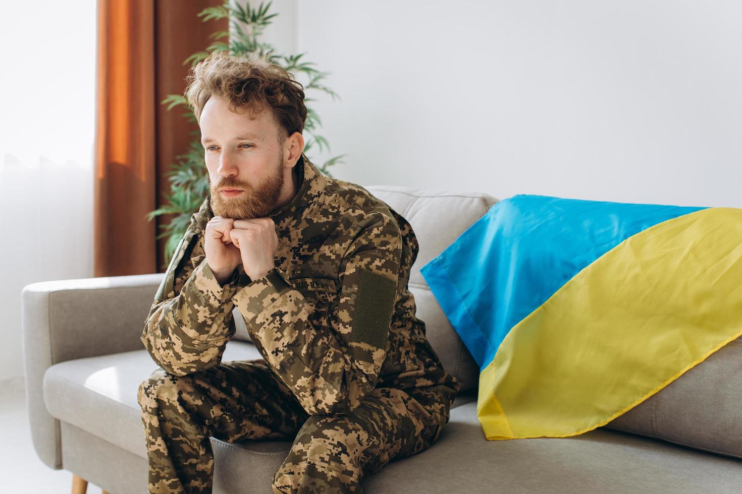 Portrait of an emotional young Ukrainian patriot soldier in military uniform sitting on the office on a sofa with a yellow and blue flag. photo