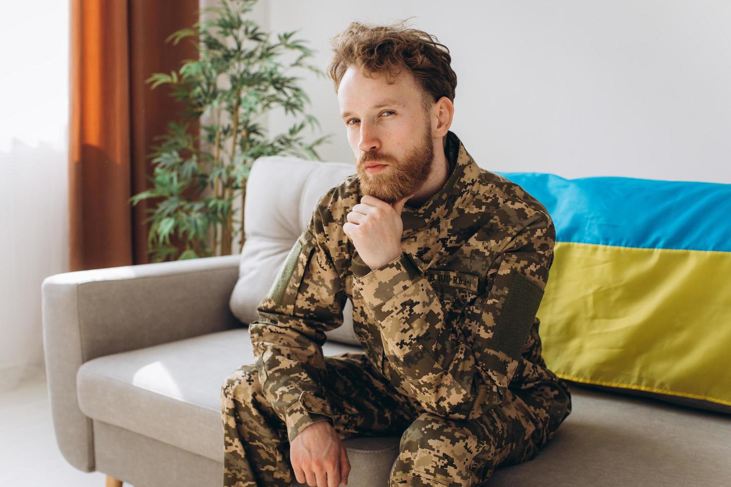 Portrait of an emotional young Ukrainian patriot soldier in military uniform sitting on the office on a sofa with a yellow and blue flag. photo