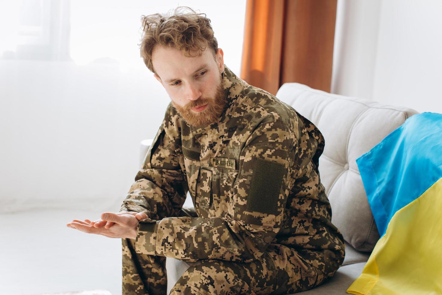 Portrait of an emotional young Ukrainian patriot soldier in military uniform sitting on the office on a sofa with a yellow and blue flag. photo