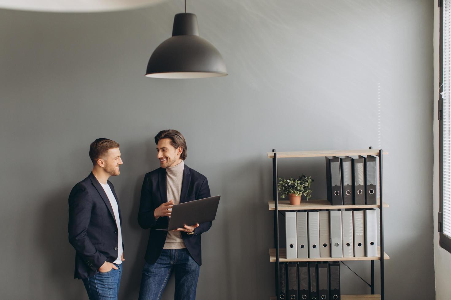 Young business men standing together holding a laptop in the modern office, discussing photo