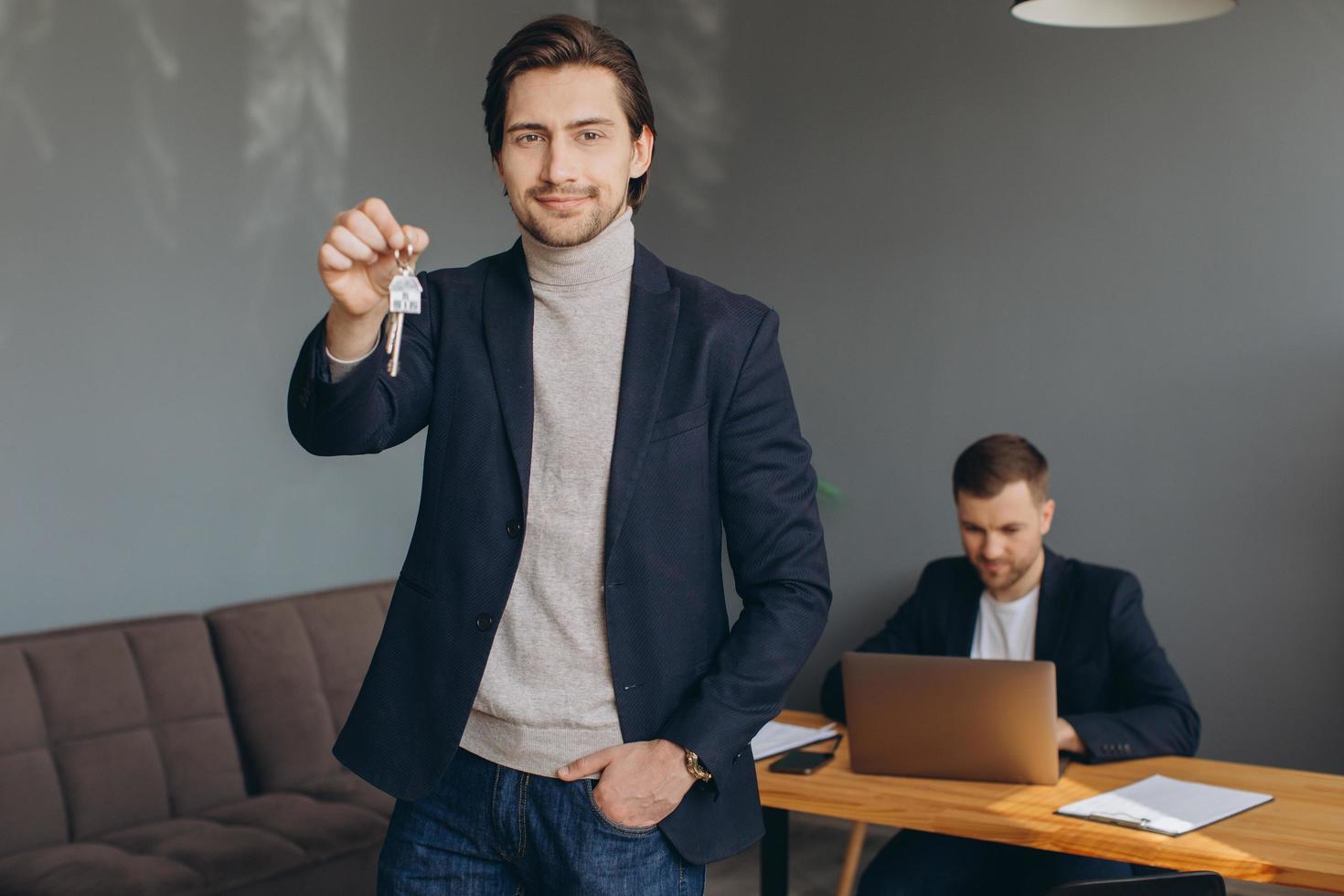 un joven moderno sonriente con traje tiene las llaves del apartamento comprado en el fondo de la inmobiliaria y la oficina foto