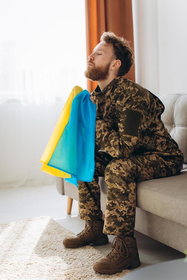 Portrait of an emotional young Ukrainian patriot soldier in military uniform sitting on the office on the couch holding a yellow and blue flag. photo