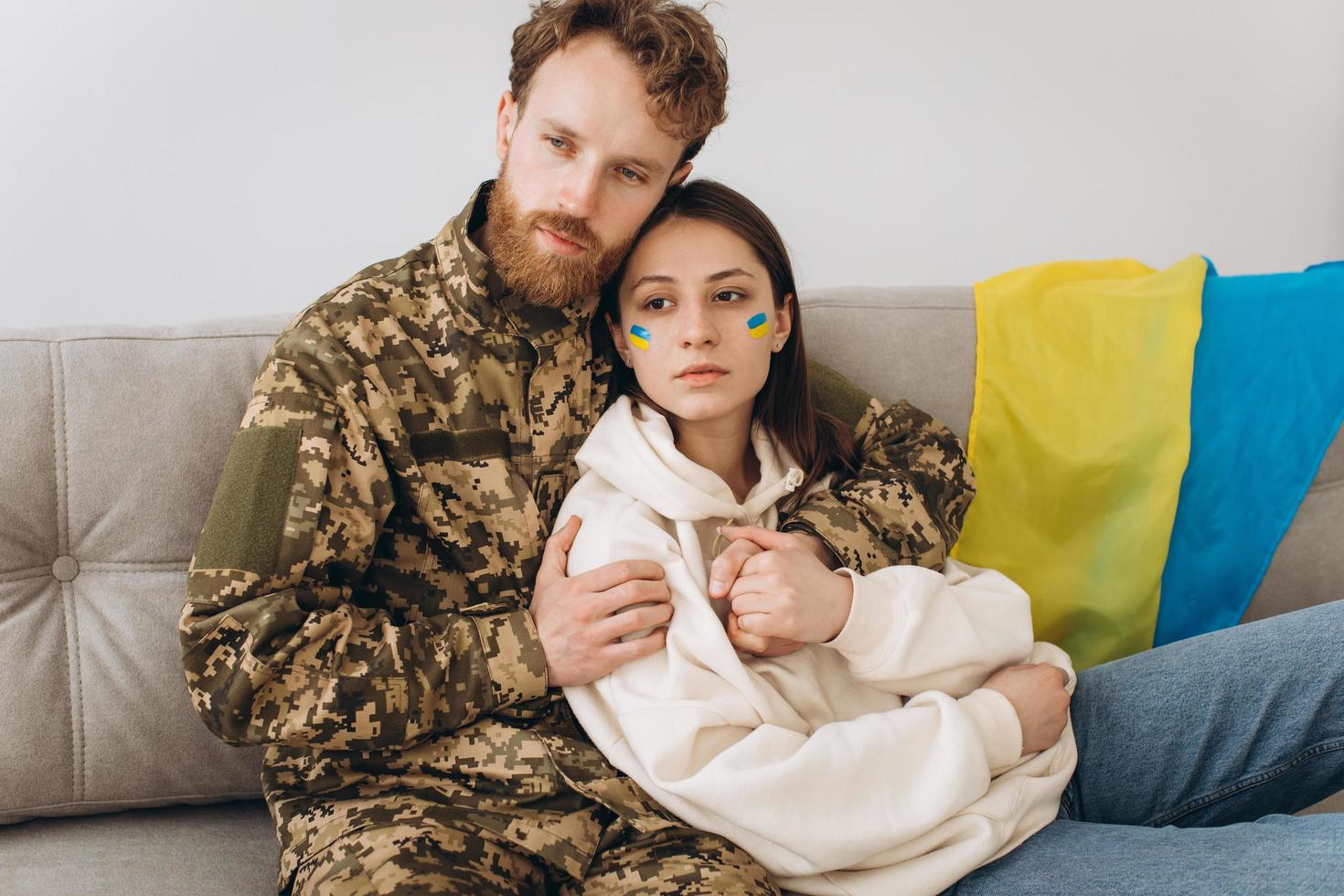Ukrainian couple, military man in uniform with his girlfriend on the couch at home on a background of yellow and blue flag photo