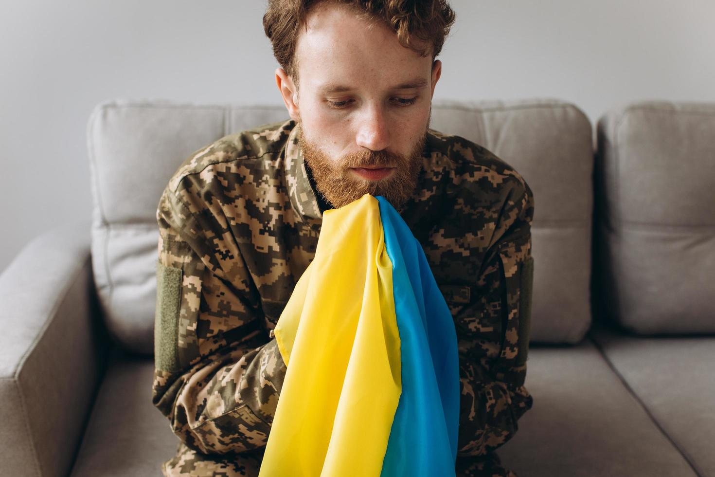 Portrait of an emotional young Ukrainian patriot soldier in military uniform sitting on the office on the couch holding a yellow and blue flag. photo