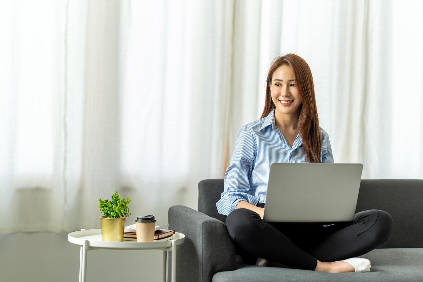 feliz mujer asiática casual y hermosa que trabaja en una computadora portátil sentada en un sofá como independiente, trabaja desde el concepto de casa. foto