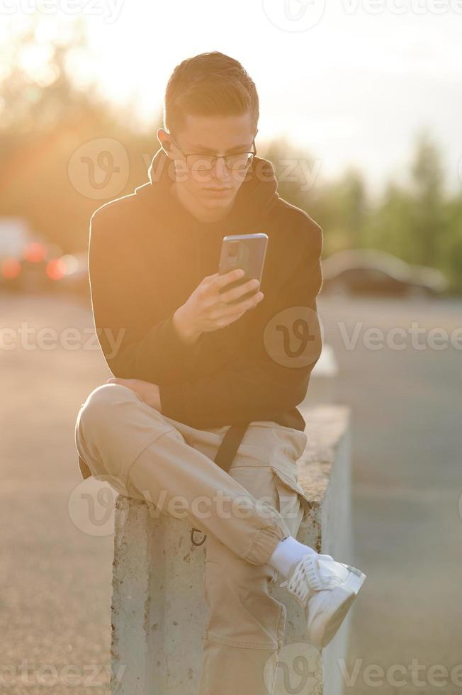 A stylish young guy in a hoodie, casual trousers and white sneakers. A guy with headphones is sitting and using a smartphone. photo