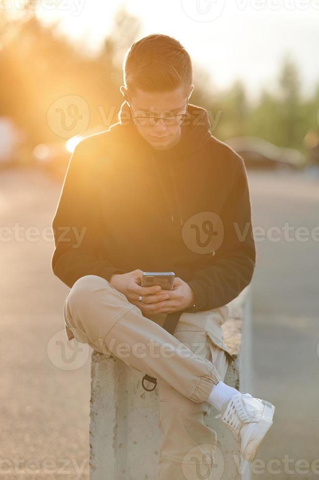 A stylish young guy in a hoodie, casual trousers and white sneakers. A guy with headphones is sitting and using a smartphone. Photo toned orange.