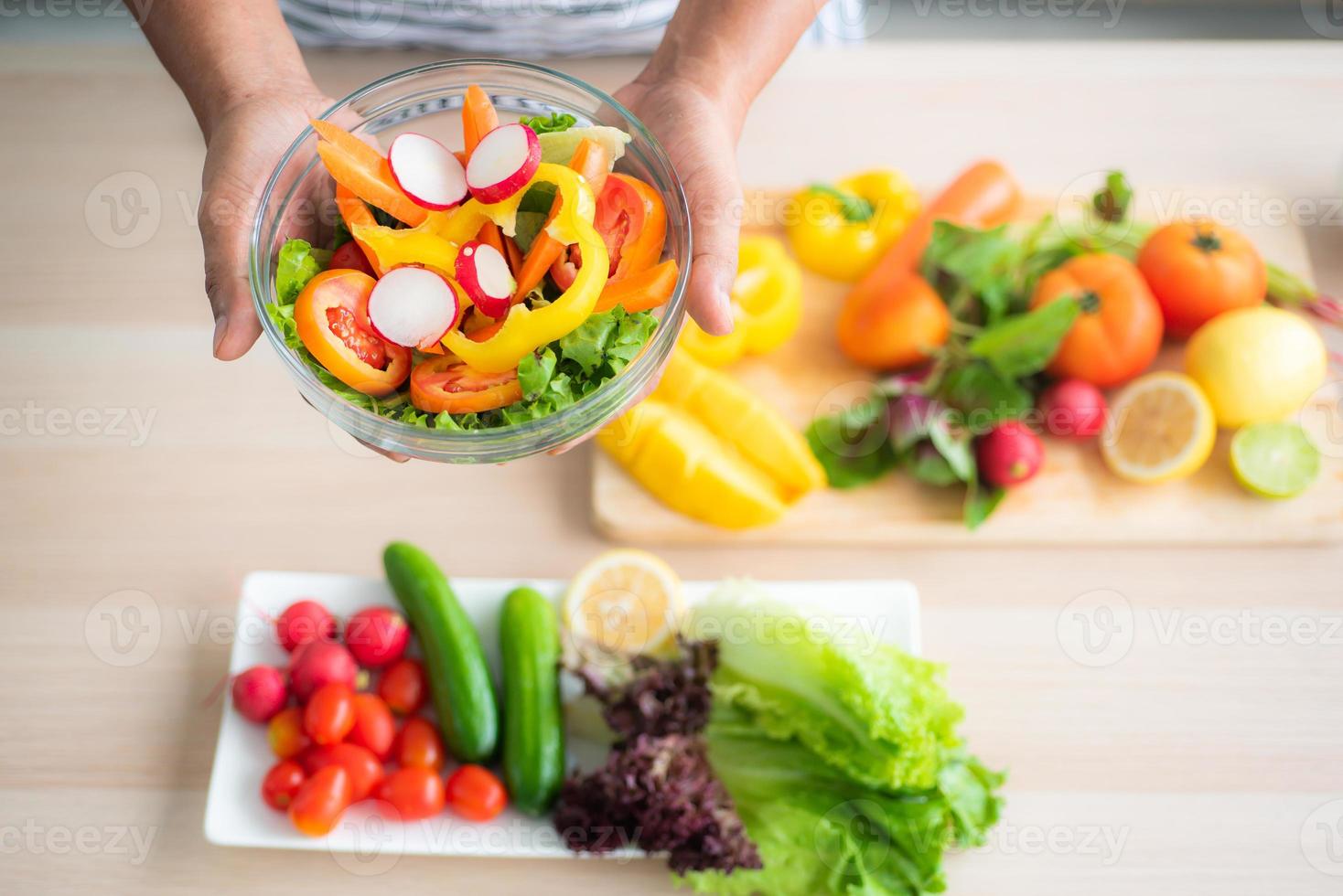 close-up top view of a vegetable salad holding in hand against a blurred background of vegetables on the table such as tomatoes, cucumbers, green oak, red oak, lemon in the kitchen. photo