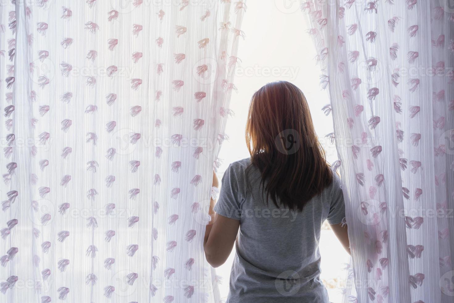 Young asian woman unveil curtain and looking out of window in the morning photo