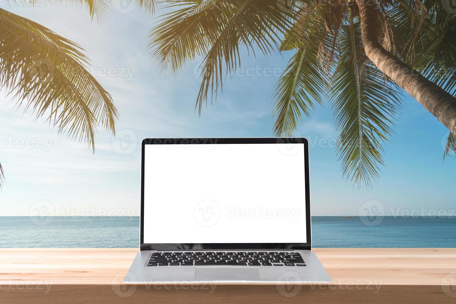 Empty screen laptop on a wooden table with coconut plam tree and sunset at tropical beach, Summer vacation concept photo