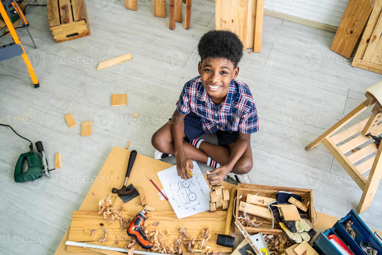 Portrait of smiling cute little boy as a construction worker working with wood sitting at floor and taking measurements making wooden model, playing with tools photo