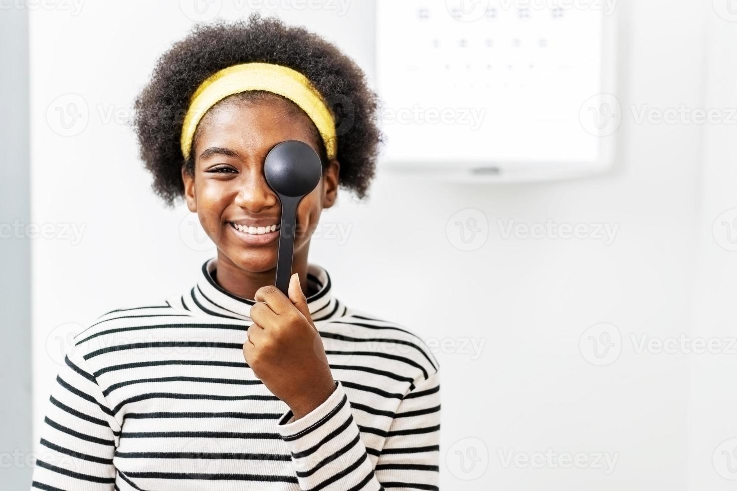Smiling young woman african american doing eye test in ophthalmological clinics, holding occluder and looking at chart, Eyesight correction, Optics clinic photo