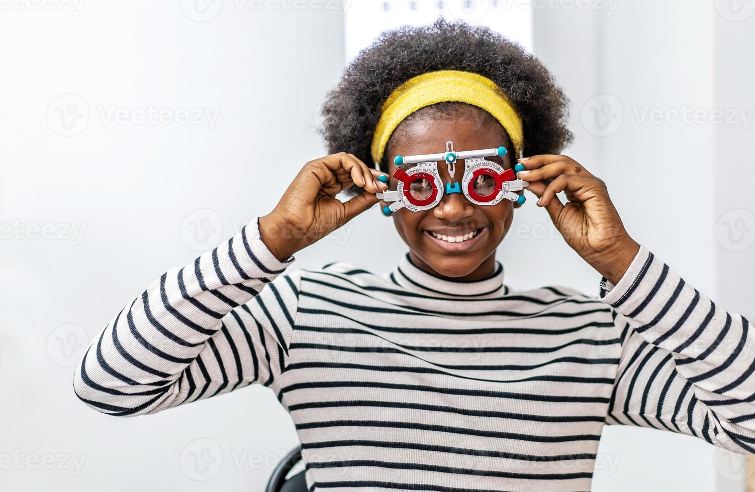 sonriente joven afroamericana revisando la visión con anteojos de prueba ocular durante un examen médico en la oficina oftalmológica, revisando la visión ocular por concepto de examen de salud óptico foto