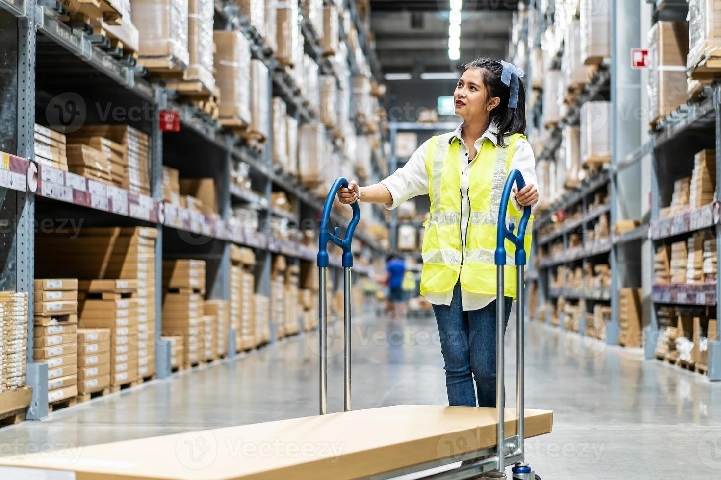 Beautiful young Asian woman staff worker pushing trolley or picking cart  to arrange things in warehouse store with blur background of boxes on shelf photo