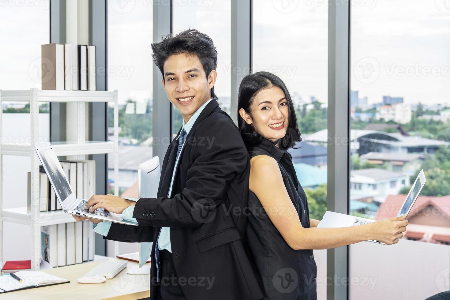 Businessman and businesswoman stand back to each other in suits smiling at the camera and working on laptops in office photo