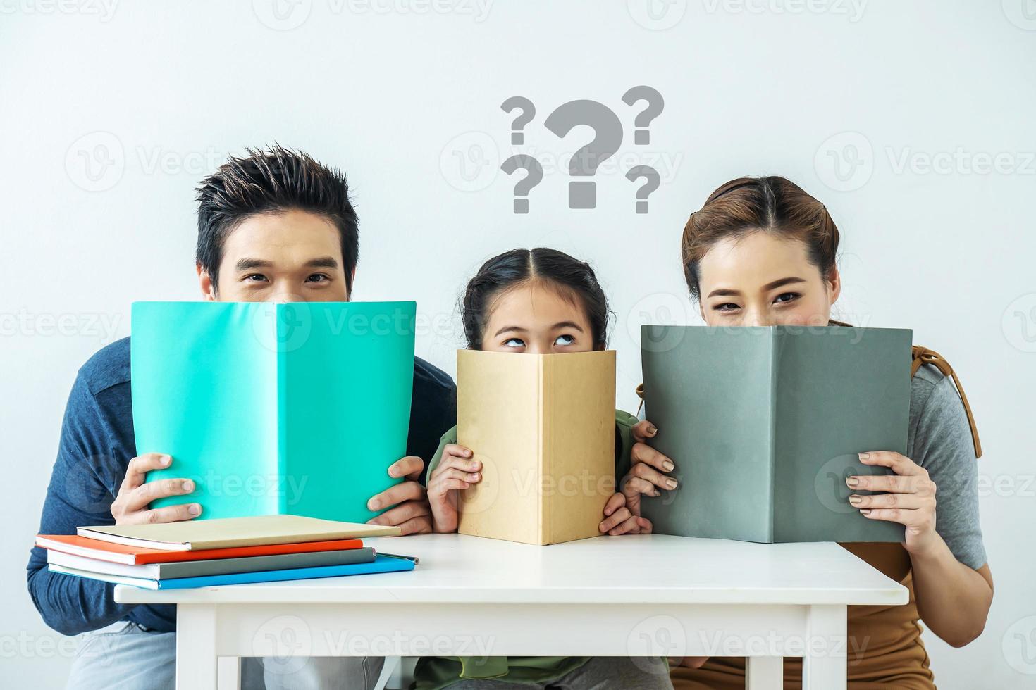 Portrait of adorable attractive beautiful asian family, Asian young mother and father with little daughter and question mark over the head sit at desk and holding book with funny facial expression photo