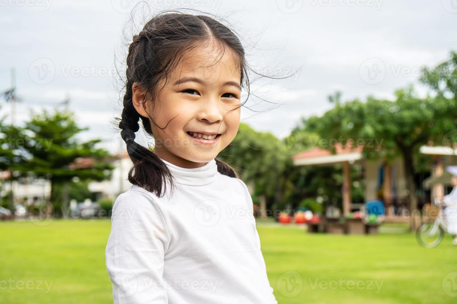 retrato de una linda niñita asiática parada en un parque de verano mirando a la cámara sonriendo alegremente, riéndose, expresivas expresiones faciales. foto