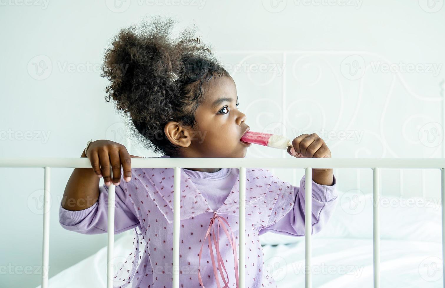 linda niña afroamericana sosteniendo comiendo helado en la cama en el dormitorio foto