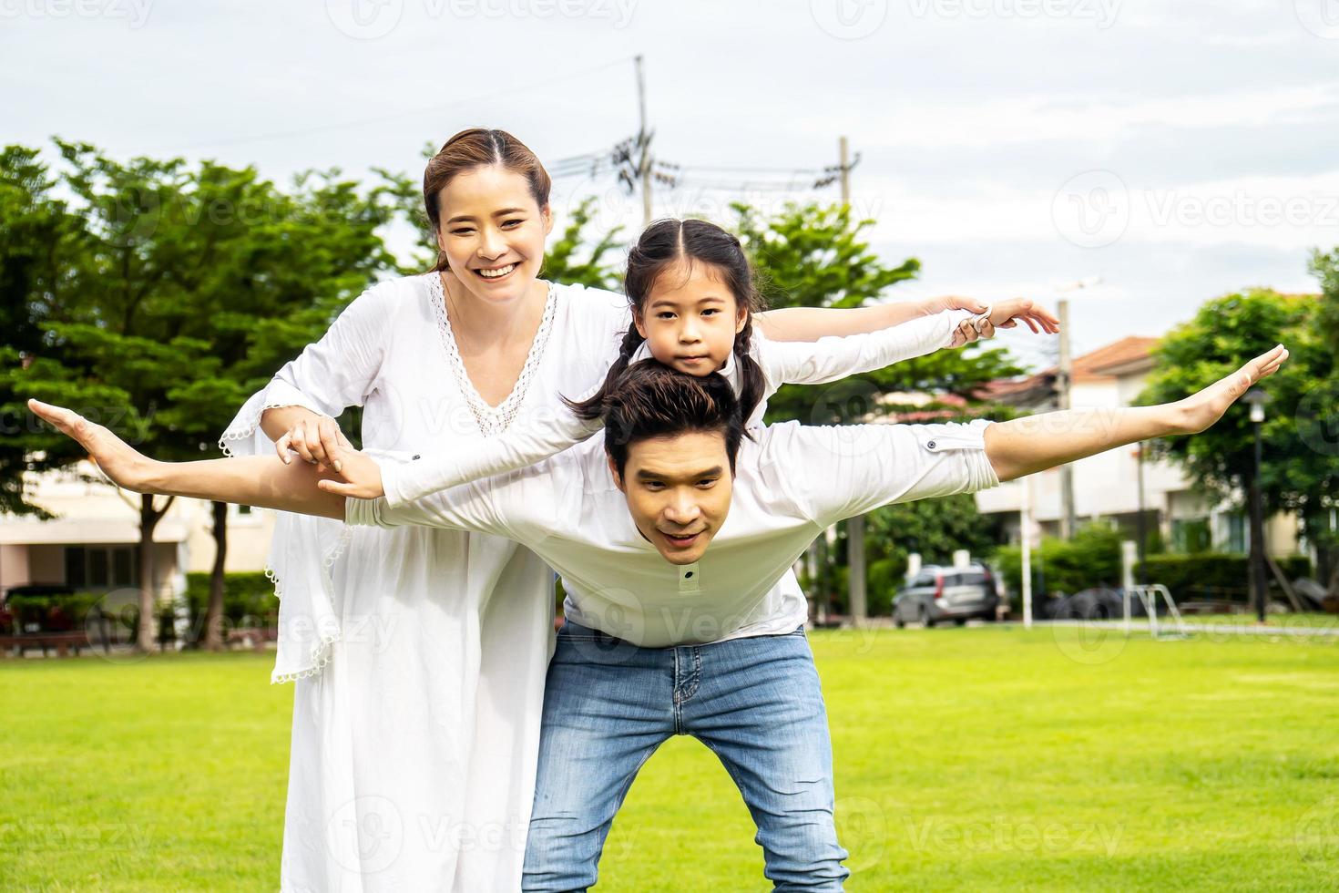 Portrait of asian happy family of three having fun together in park, little daughter sitting on father back and mother smile walking and having fun moments good time in summer park at home photo