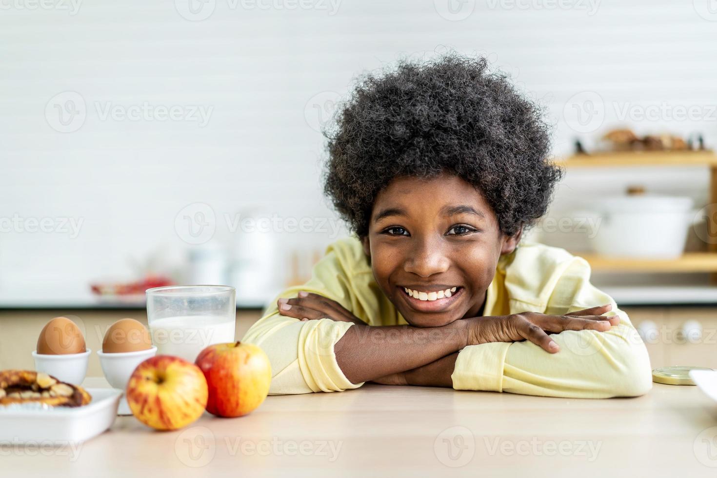 cerrar un niño sonriente bebiendo un vaso de leche, sentado en una mesa de madera en la cocina, un niño adorable disfrutando de yogur orgánico, recibiendo vitaminas y calcio, concepto de salud infantil foto
