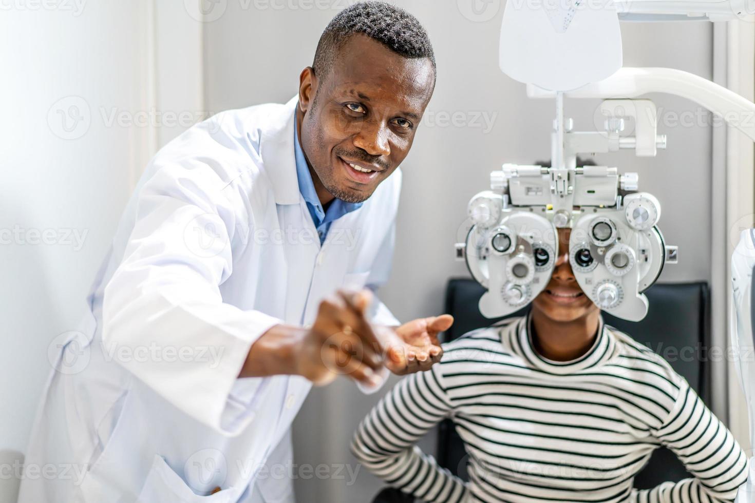 Optician doing optometry eye exam for black african american teen girl patient. Male optometrist with phoropter while examining patient. photo