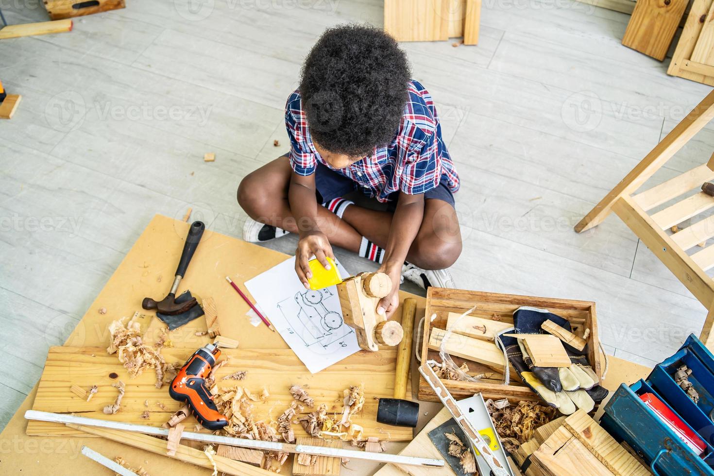 niño lindo como trabajador de la construcción trabajando con madera sentado en el suelo y tomando medidas haciendo modelos de madera, jugando con herramientas foto