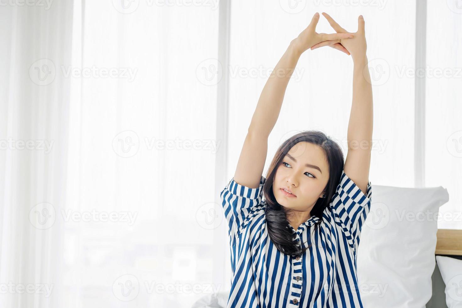 Beautiful young asian woman sitting on her bed in morning, stretching with arms raised after waking up, happy and relaxed after good night sleep,Healthy lifestyle, Wellness concept photo