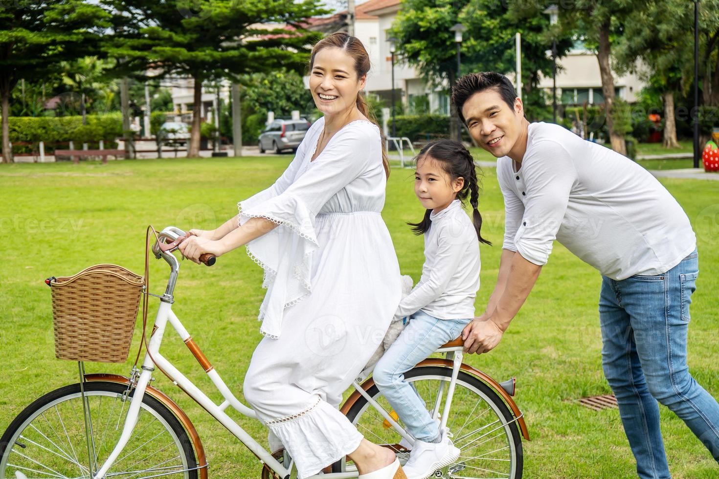 Happy family with bicycle outdoors on summer day, Asian father and mother with daughter enjoying riding a bike outdoors in a park. photo