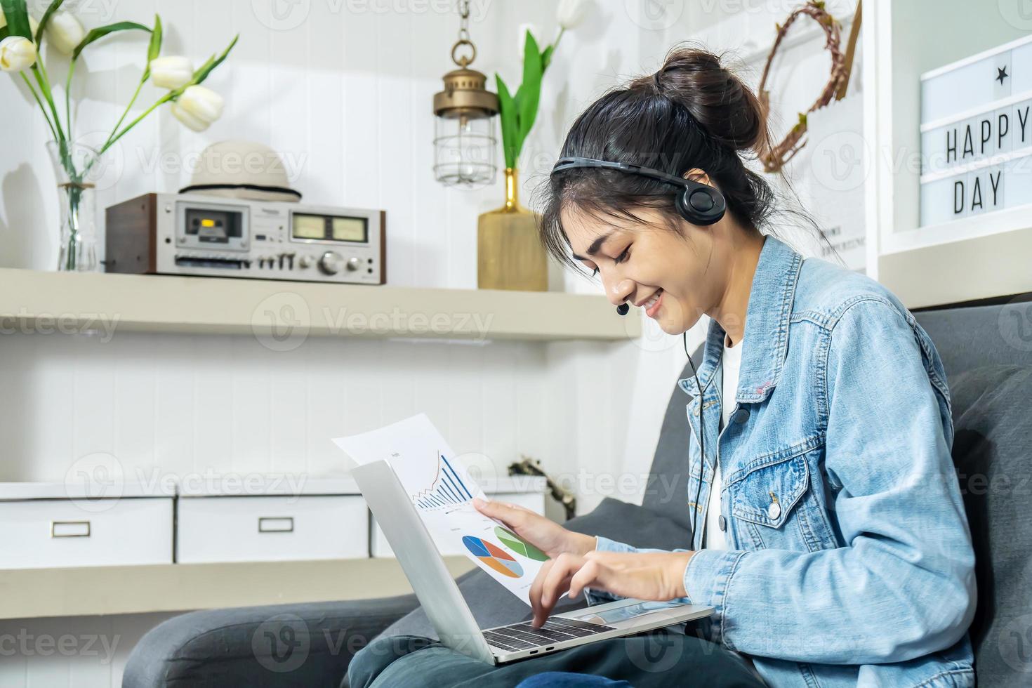 Asian young business woman is sitting on the sofa in her own house is wearing headphones video call conference with laptop computer with pile of graph and chart paper at home office, work from home photo
