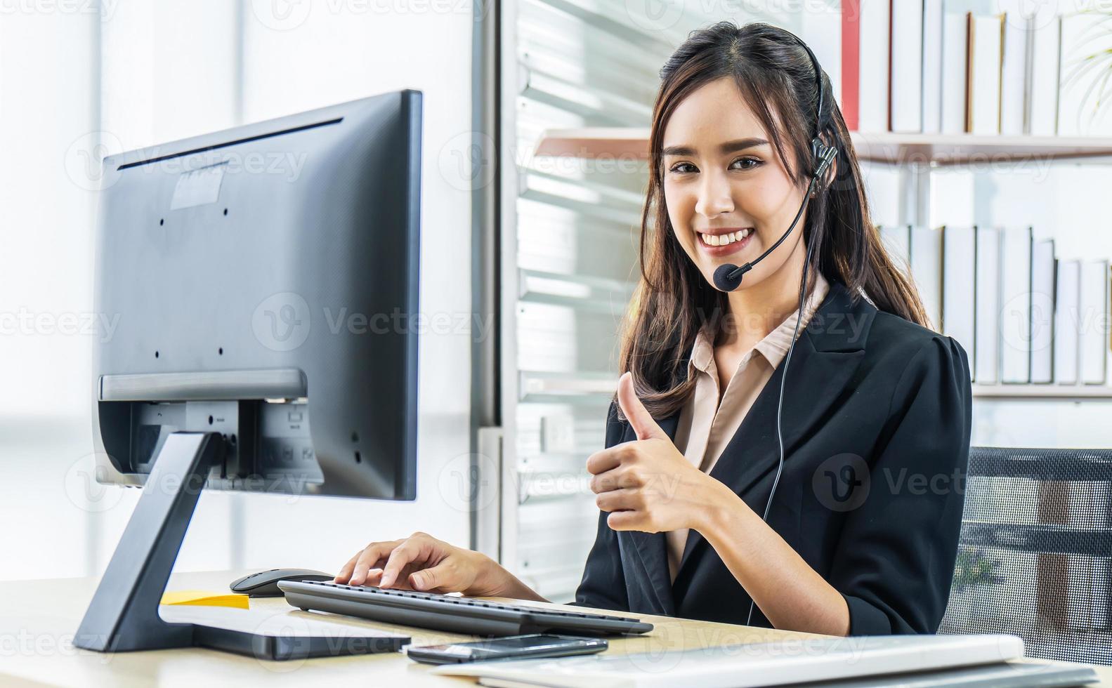 sonriente y amable agente asiática del centro de llamadas con auriculares que muestran su aprobación por trabajar en la línea directa de soporte en la oficina, concepto de mente de servicio foto