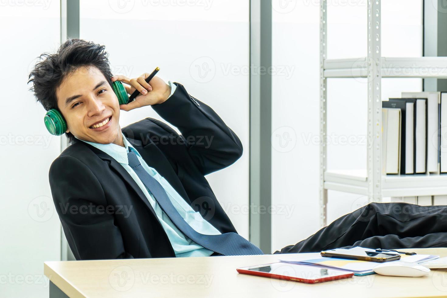 Happy businessman using headphones listen to music during break at workplace. employee male enjoying favorite track, listening to music throwing legs on the table. photo