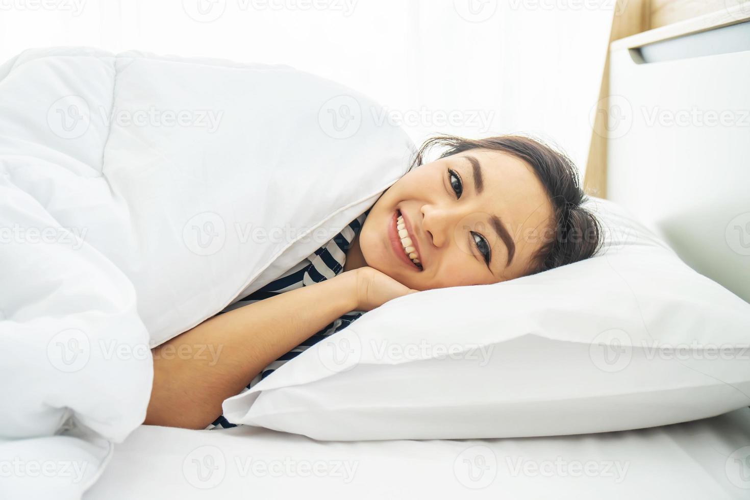 Young beautiful woman smiling while lying on white pillow in bed at early morning photo