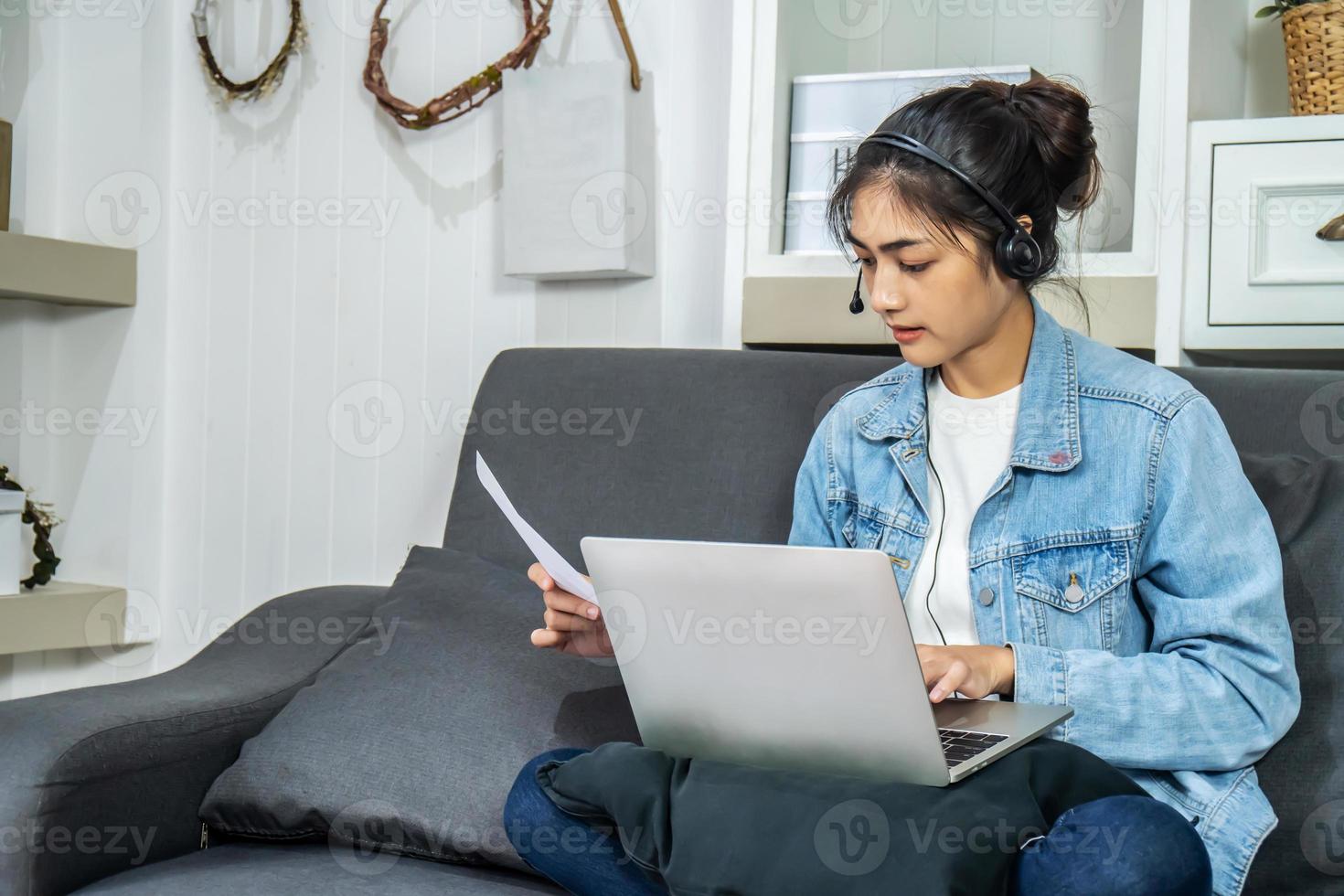 Asian young business woman is sitting on the sofa in her own house is wearing headphones video call conference with laptop computer with pile of graph and chart paper at home office, work from home photo