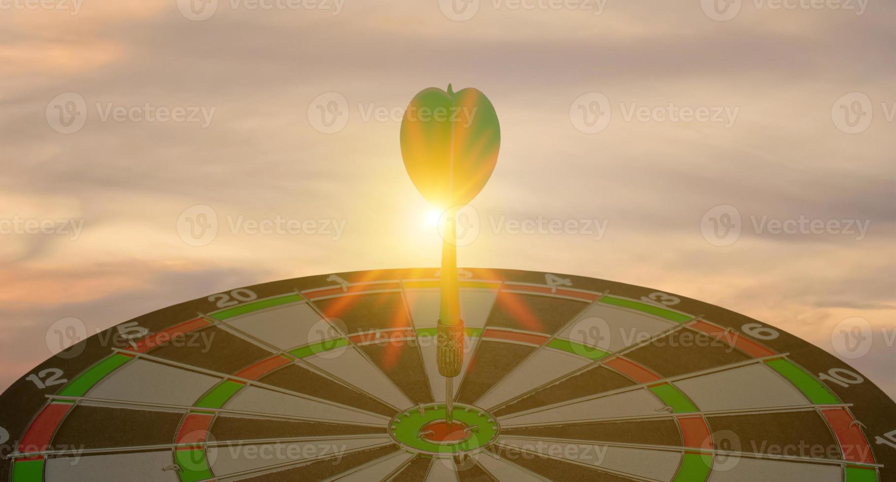 silueta de flecha de dardo verde que golpea el tablero de dardos del centro de la diana en el fondo de la puesta de sol. concepto de orientación y enfoque empresarial, estrategia, logro y concepto de planificación foto