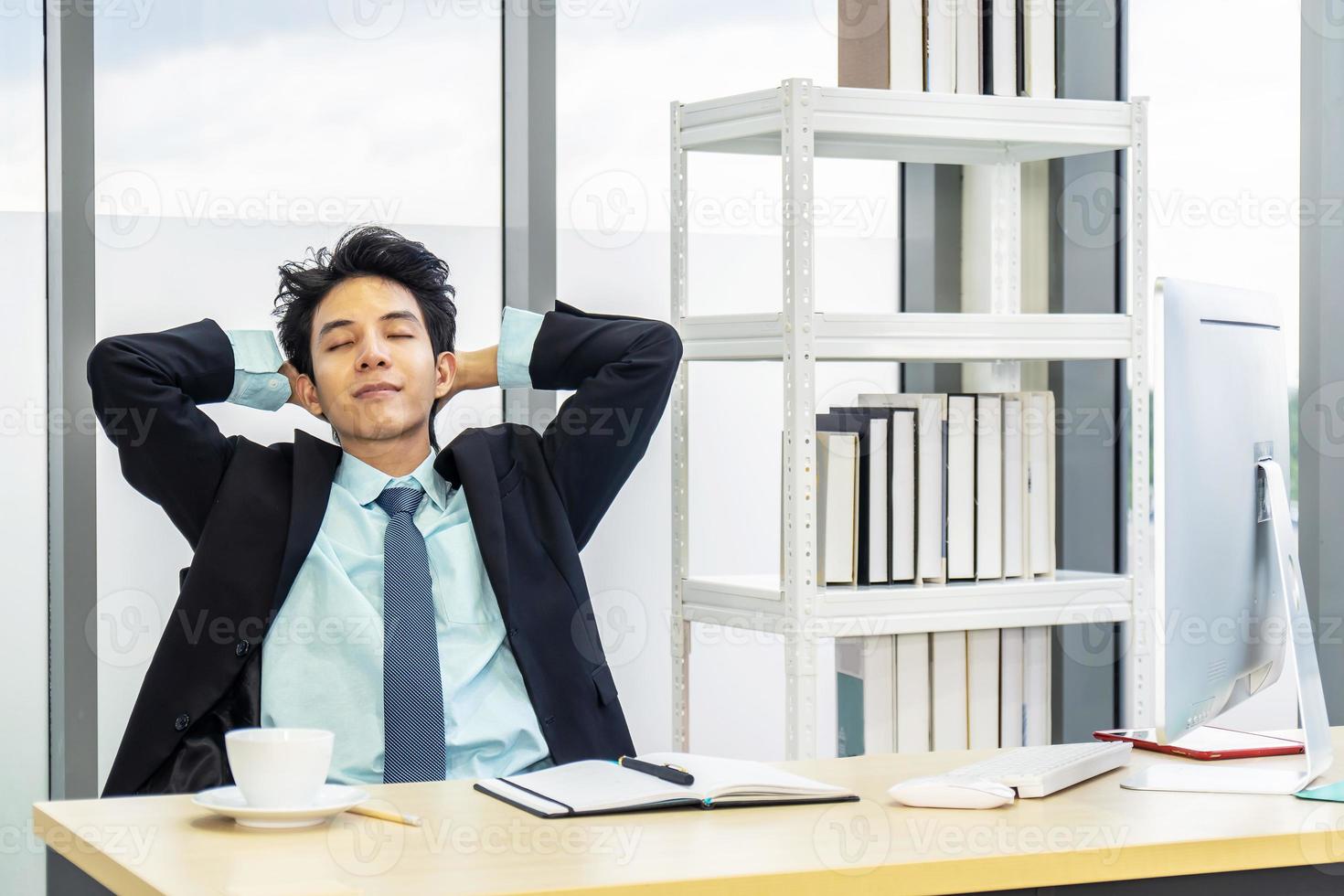 Young asian businessman having break and resting holding hands behind head and keeping eyes closed while sitting at his working place photo