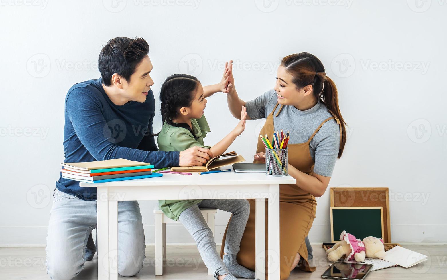 Asian young mother and father with little daughter sit at desk learning and writing in book with pencil making homework at home.Education concept. photo