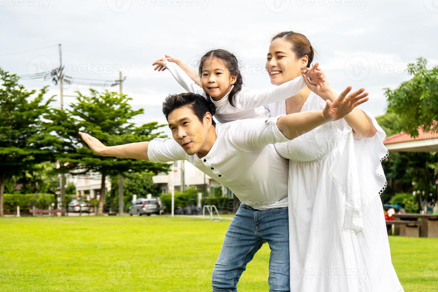 Portrait of asian happy family of three having fun together in park, little daughter sitting on father back and mother smile walking and having fun moments good time in summer park at home photo