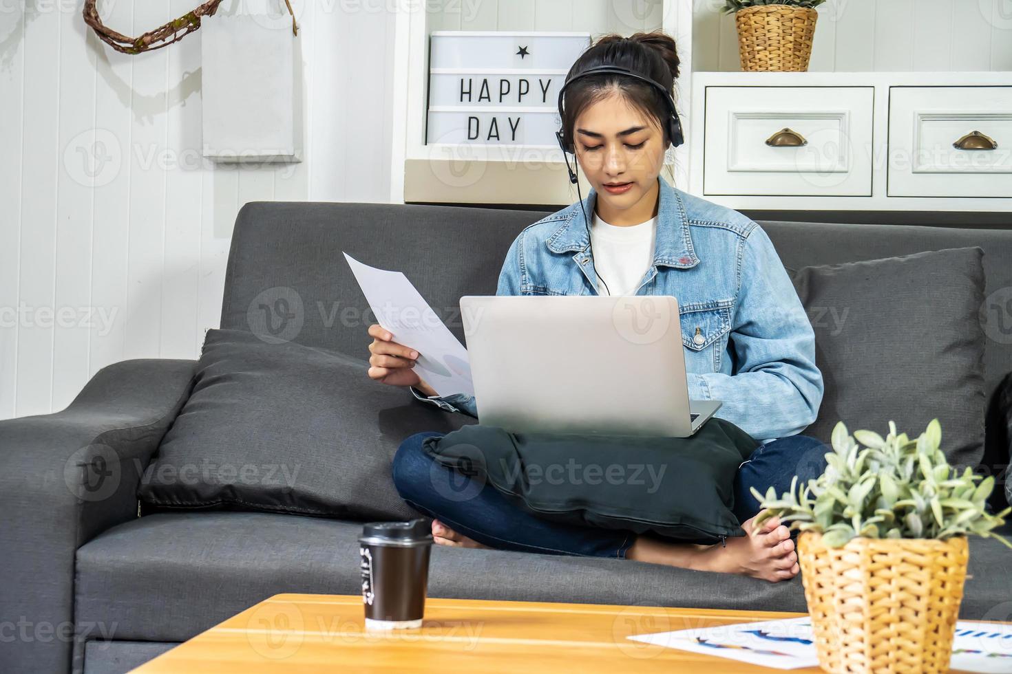 Asian young business woman is sitting on the sofa in her own house is wearing headphones video call conference with laptop computer with pile of graph and chart paper at home office, work from home photo