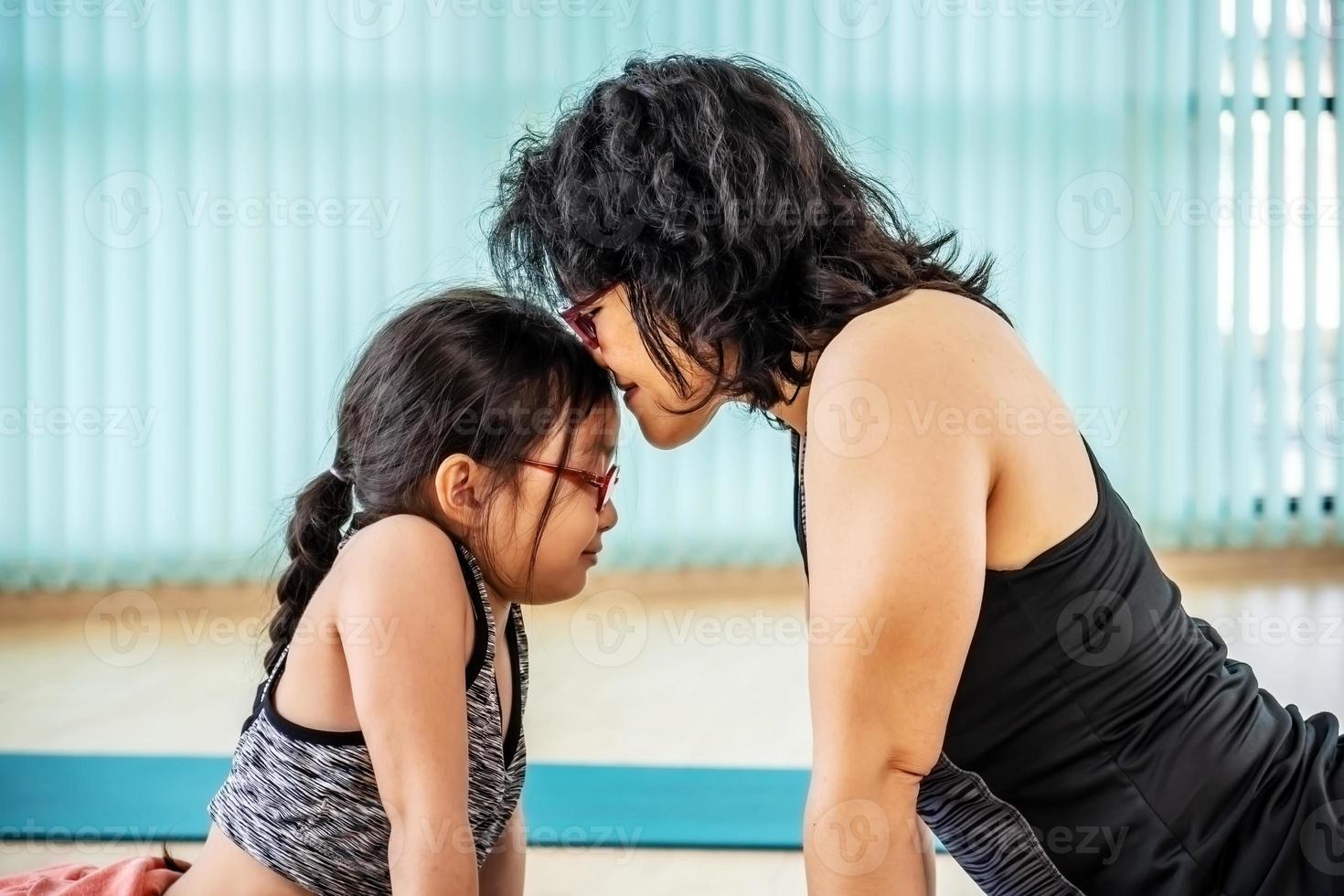 Asian fit mother with little child daughter are smiling while practicing yoga together at home, exercise class on asana mat at home. parent accompany healthy children sport concept photo