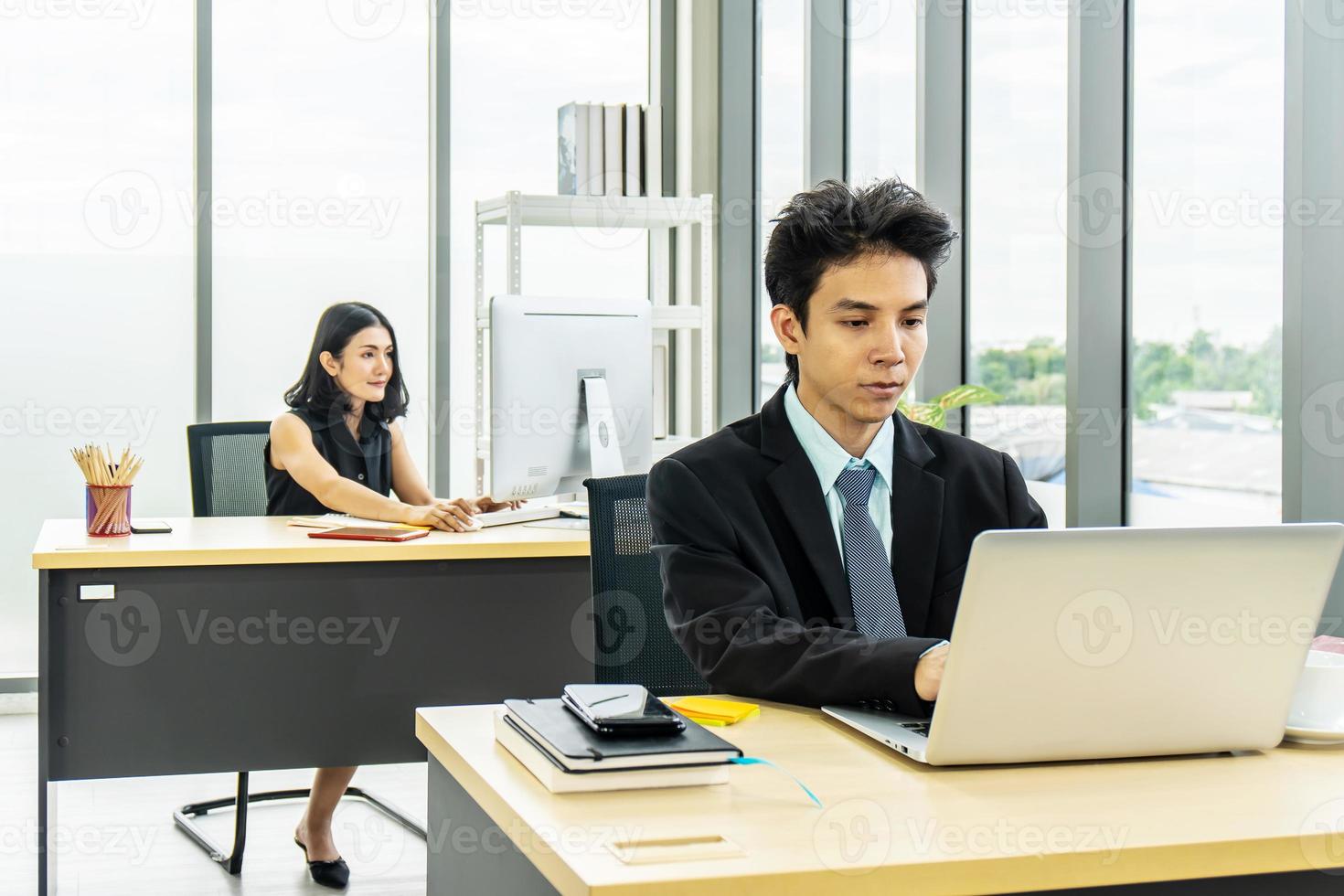 Young asian businessman sitting at desk working on laptop and businesswoman working on laptop in her workstation in background. photo