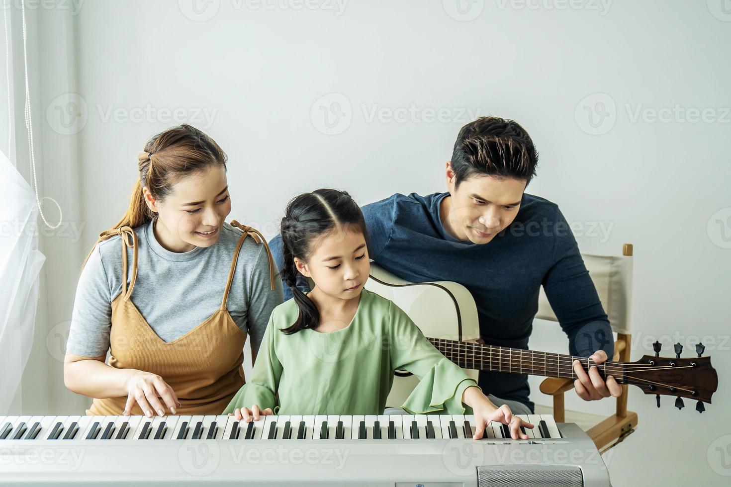 Happy little Asian daughter playing piano with mother and father play guitar at home, Mother teaching daughter to play piano,They play and sing songs. They are having fun. photo