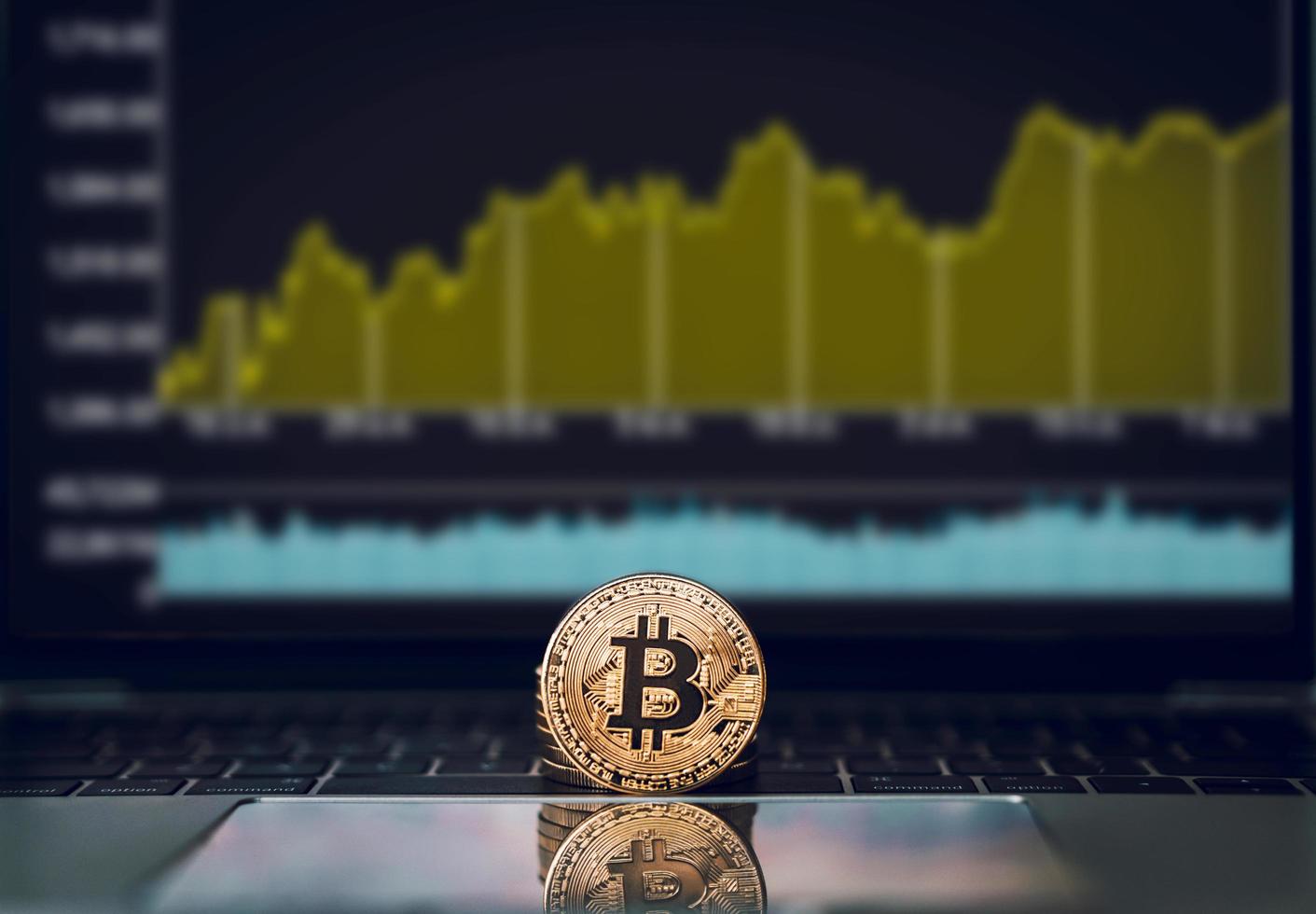 A golden bitcoin is place on the keyboard in front of a screen of stock or cryptocurrency market candles or charts or graphs with a laptop, cryptocurrency trading and investment concept photo